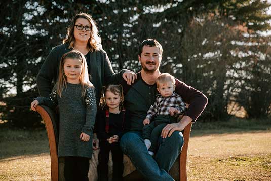 Brady and Lacey Merriam with their 3 children sitting on a bench in front of pine trees
