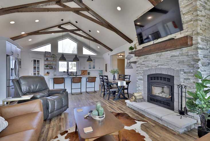 View from open living room to kitchen featuring vaulted ceiling, white cabinets and light-colored stacked stone fireplace