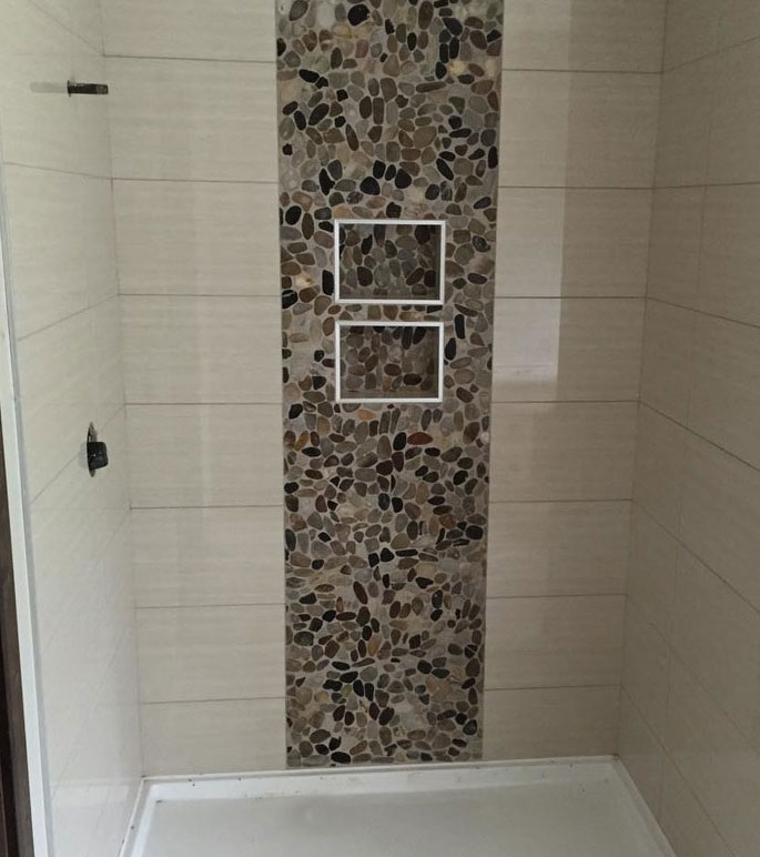 Large cream tile in shower stall is accented by wide pebble tile panel with 2 shampoo niches